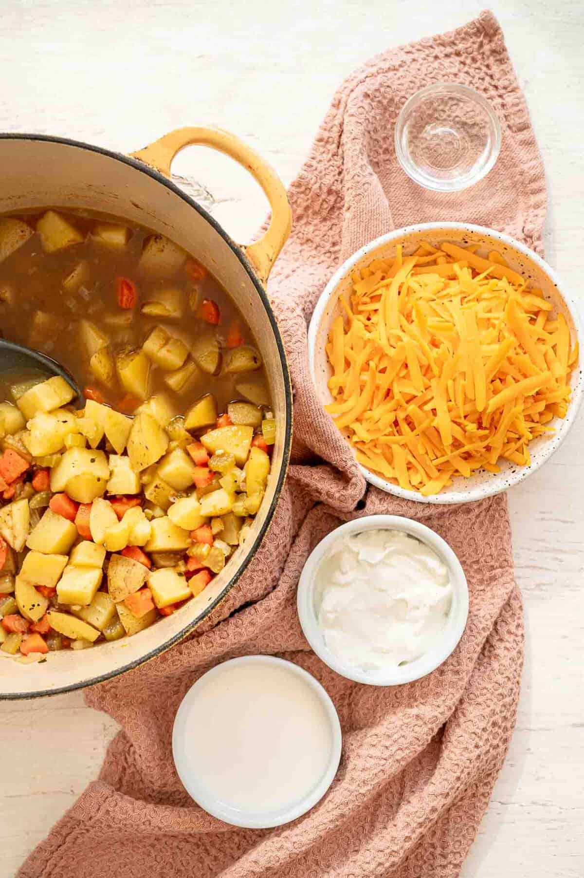 Potato soup ingredients in a pot with bowls of half and half, sour cream, cheddar cheese, and white vinegar next to it.