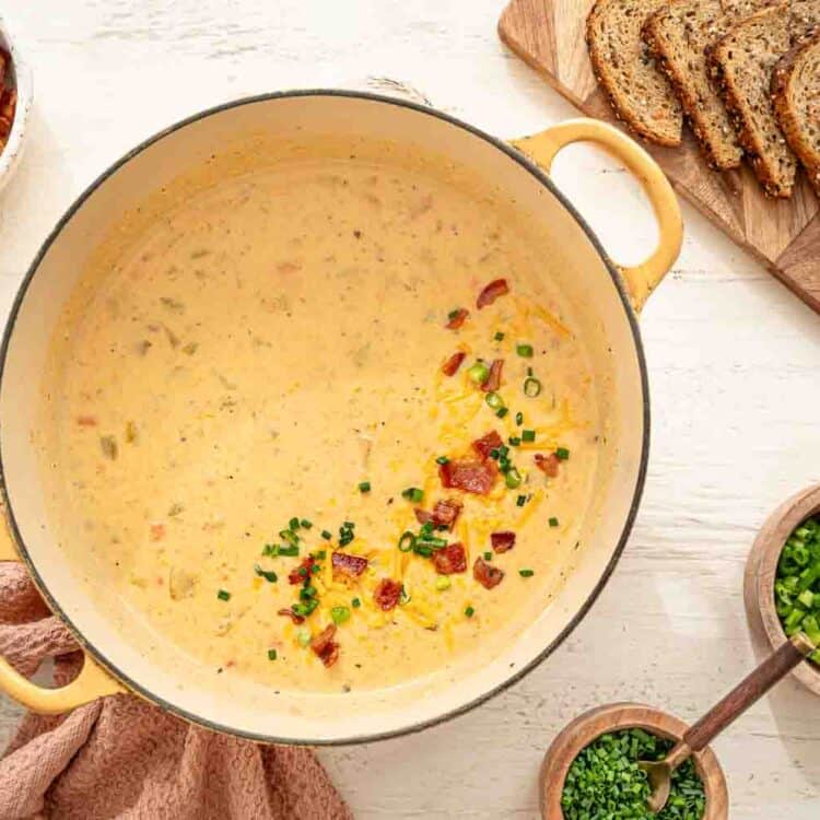 Cheddar and bacon soup in a dutch oven with chopped bacon, and green onions in bowls next to it.