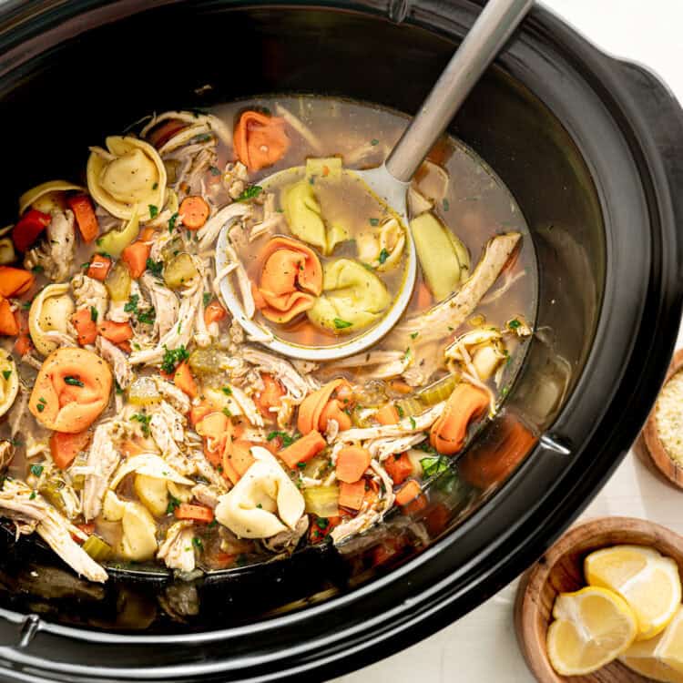 Chicken tortellini in a slow cooker with small bowls of Parmesan cheese and lemon wedges next to it.