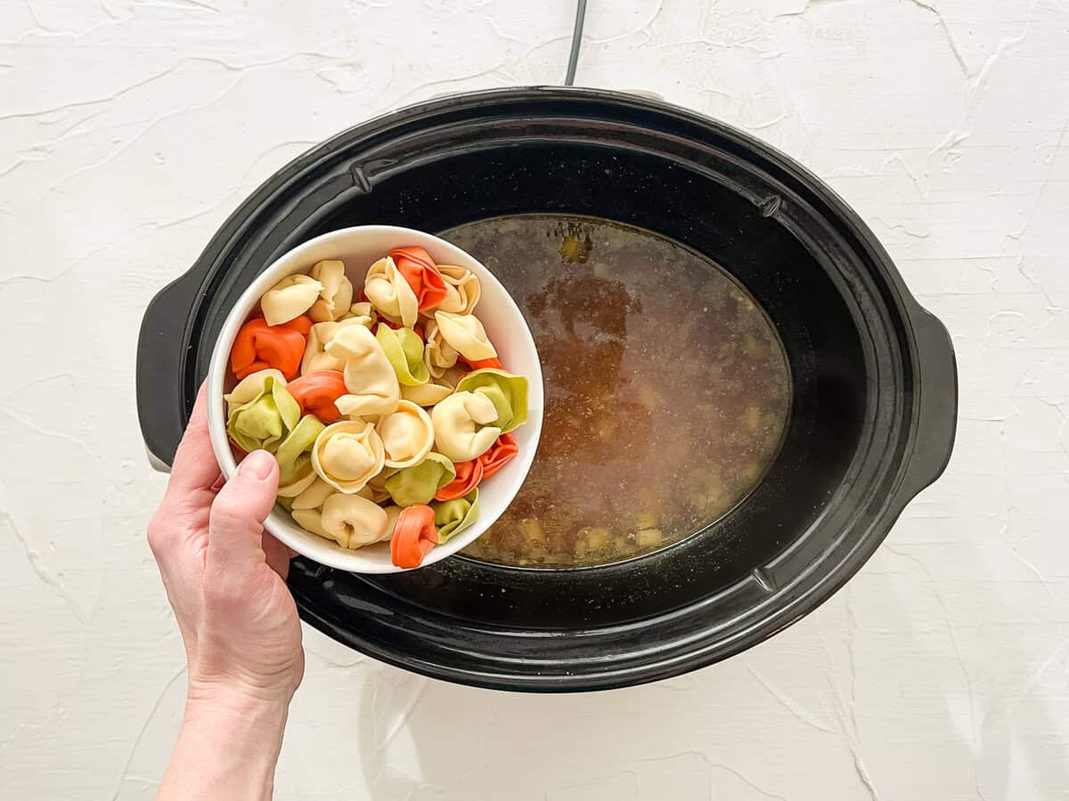 Cheese tortellini being dumped into a slow cooker with the rest of the ingredients for chicken tortellini soup.