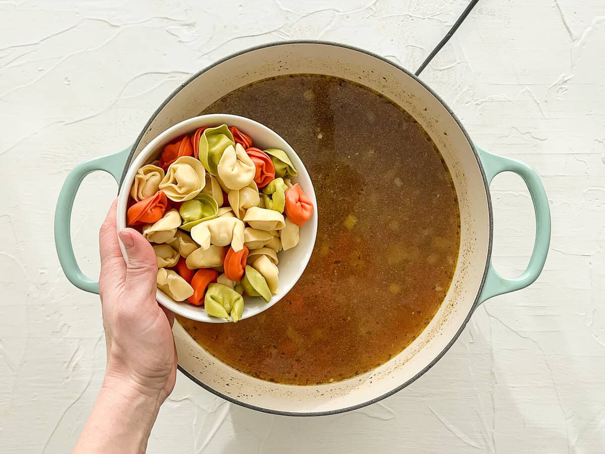 Cheese tortellini being dumped into a pot of broth and other ingredients.