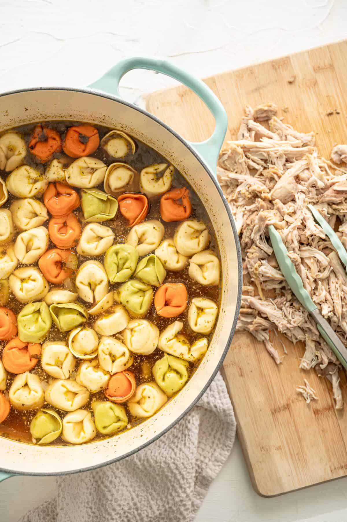 Large pot with cheese tortellini in soup broth, and a wooden cutting board with shredded chicken next to it.