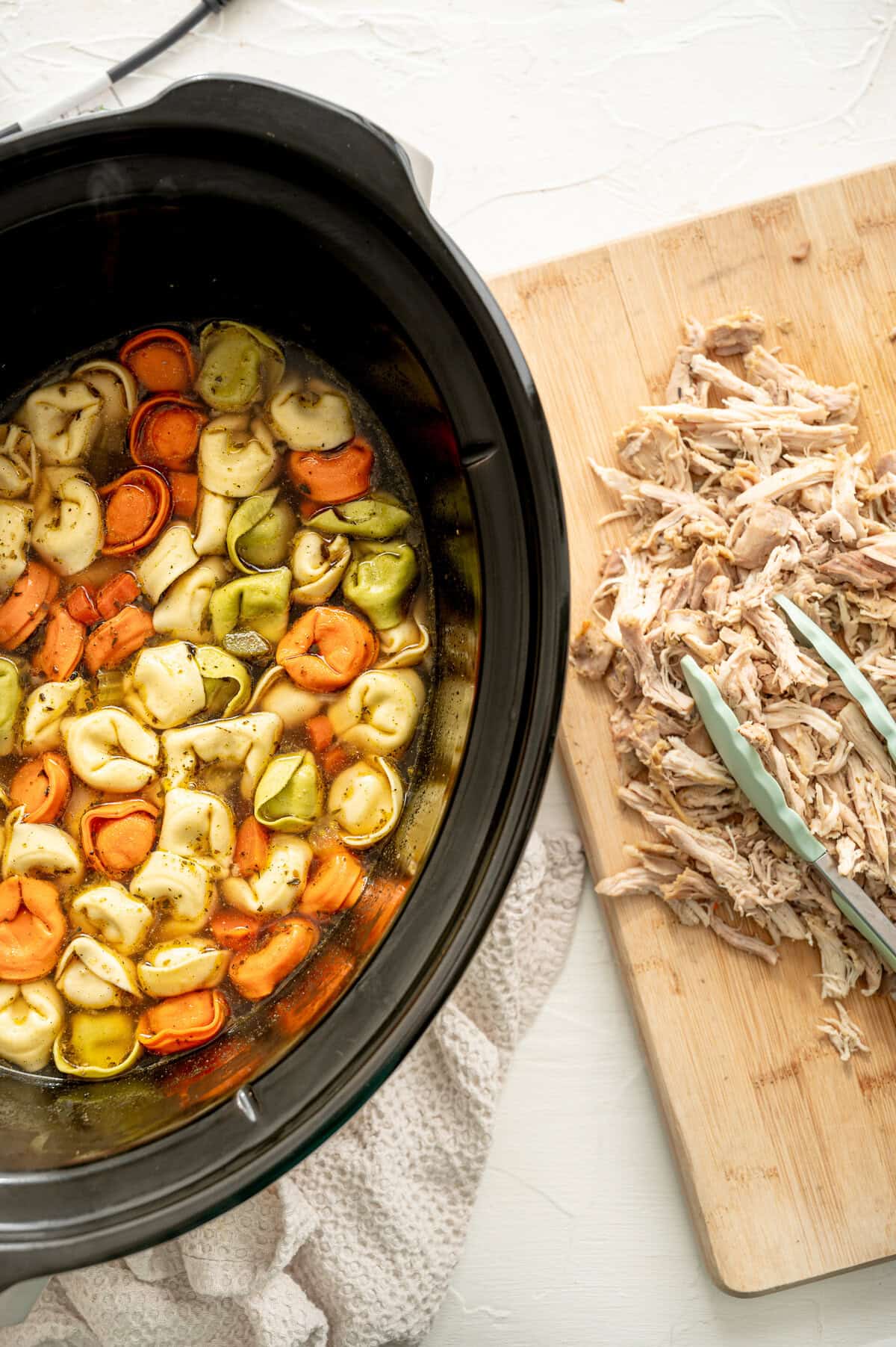 Slow cooker with cheese tortellini in soup broth, and a wooden cutting board with shredded chicken next to it.