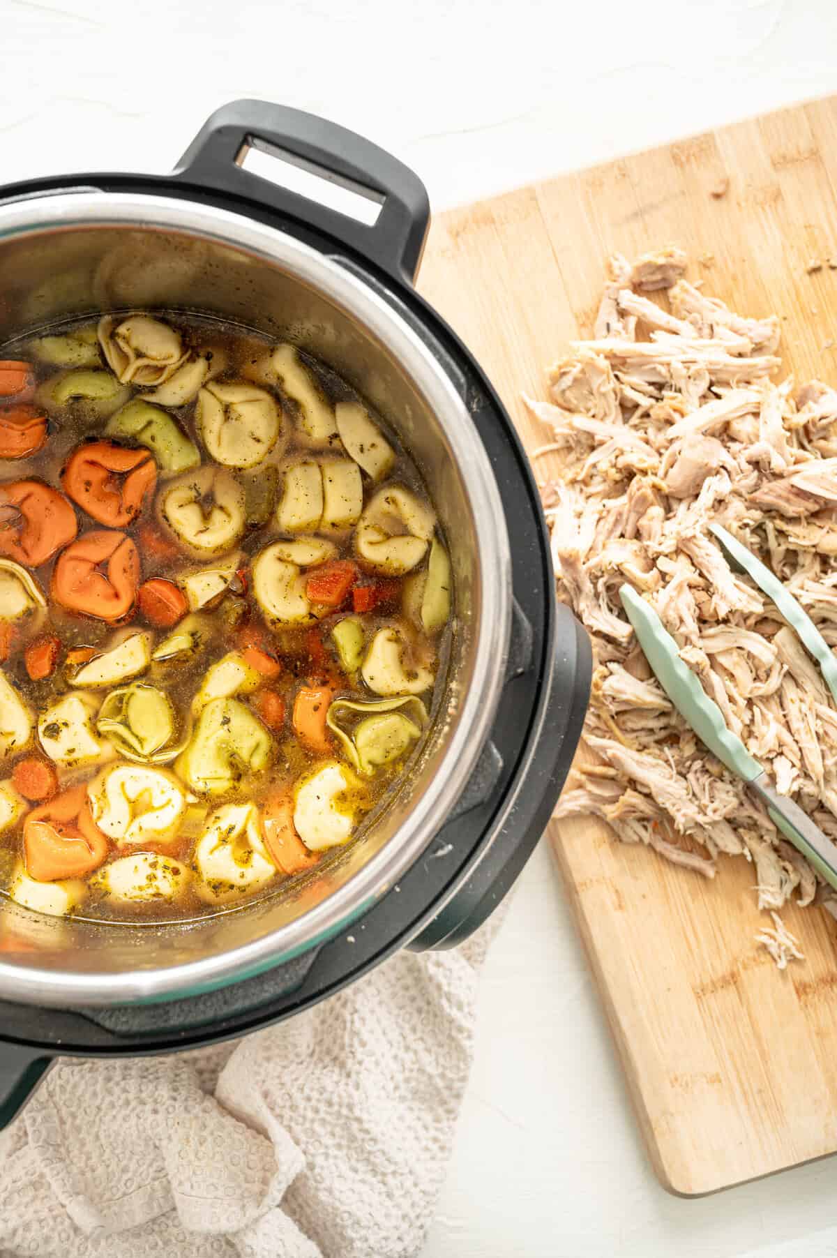 Instant Pot with cheese tortellini in soup broth, and a wooden cutting board with shredded chicken next to it.