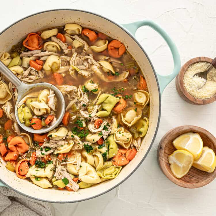 Chicken tortellini soup in a pot with small bowls of Parmesan cheese and lemon wedges.