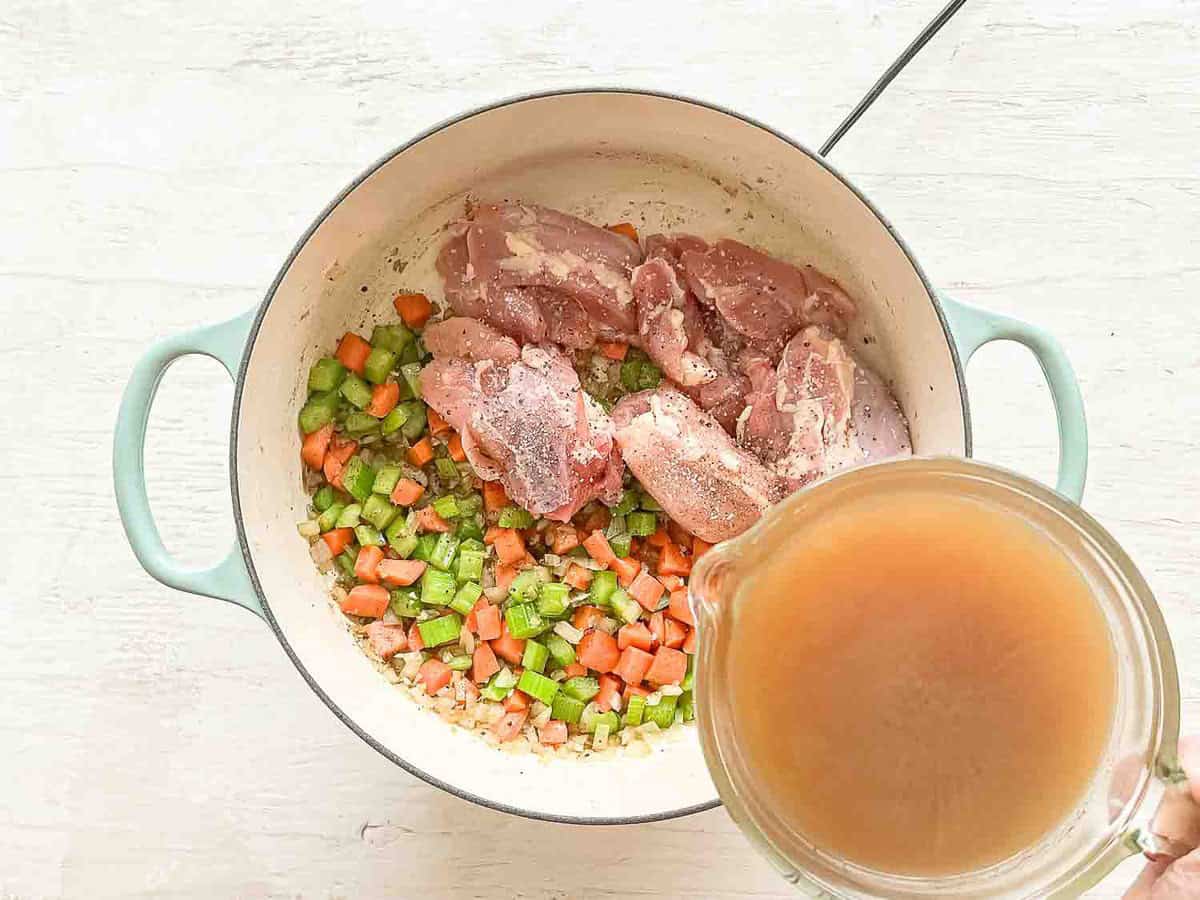 Chicken broth being poured into a pot with sauted veggies and raw boneless, skinless chicken thighs.