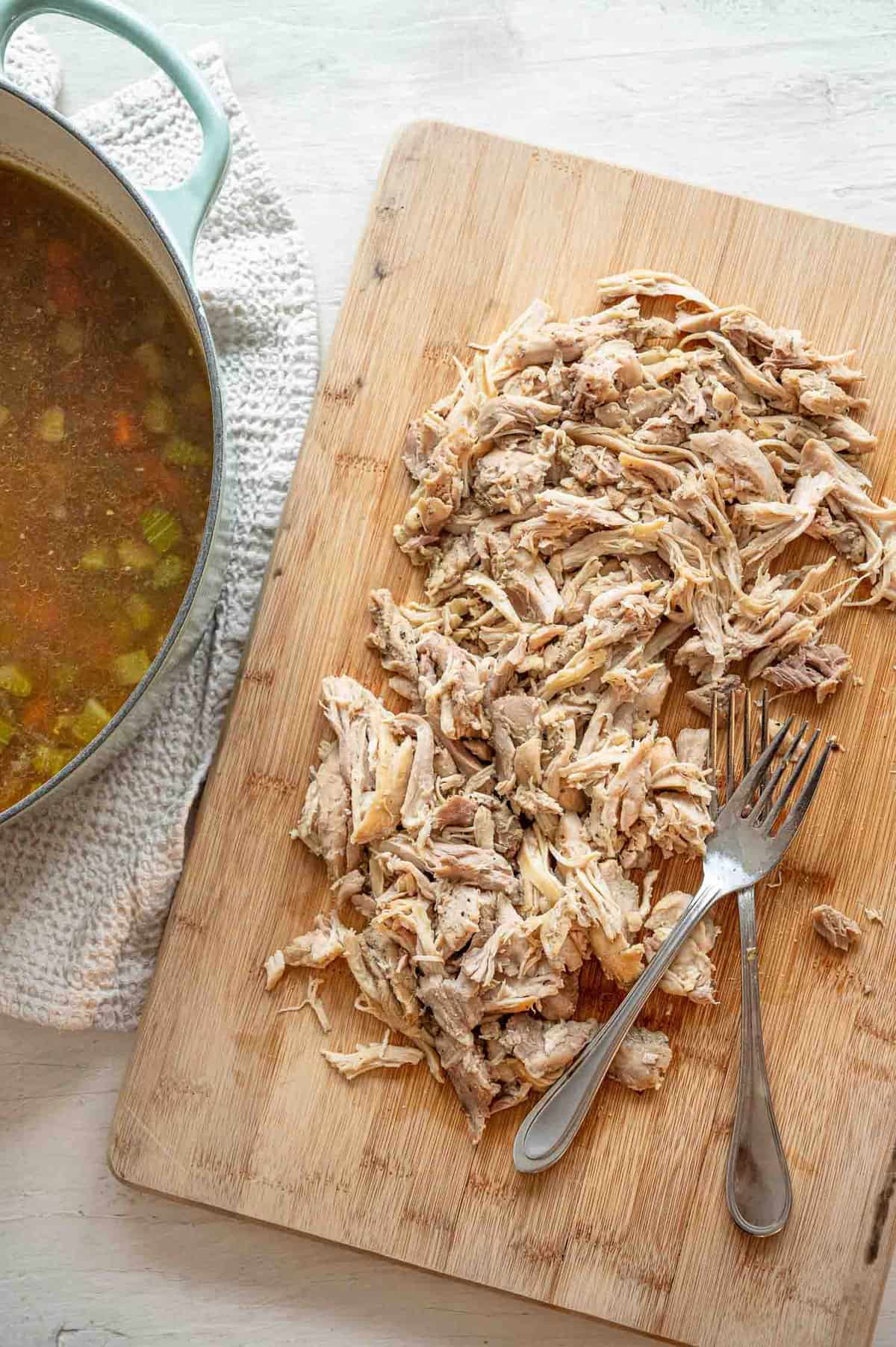 Shredded chicken and two forks on a wooden cutting board next to a pot with broth and veggies.