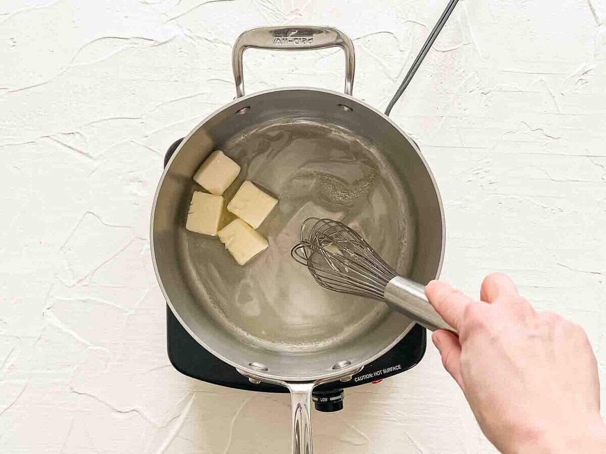 A hand whisking butter in a saucepan as it melts.