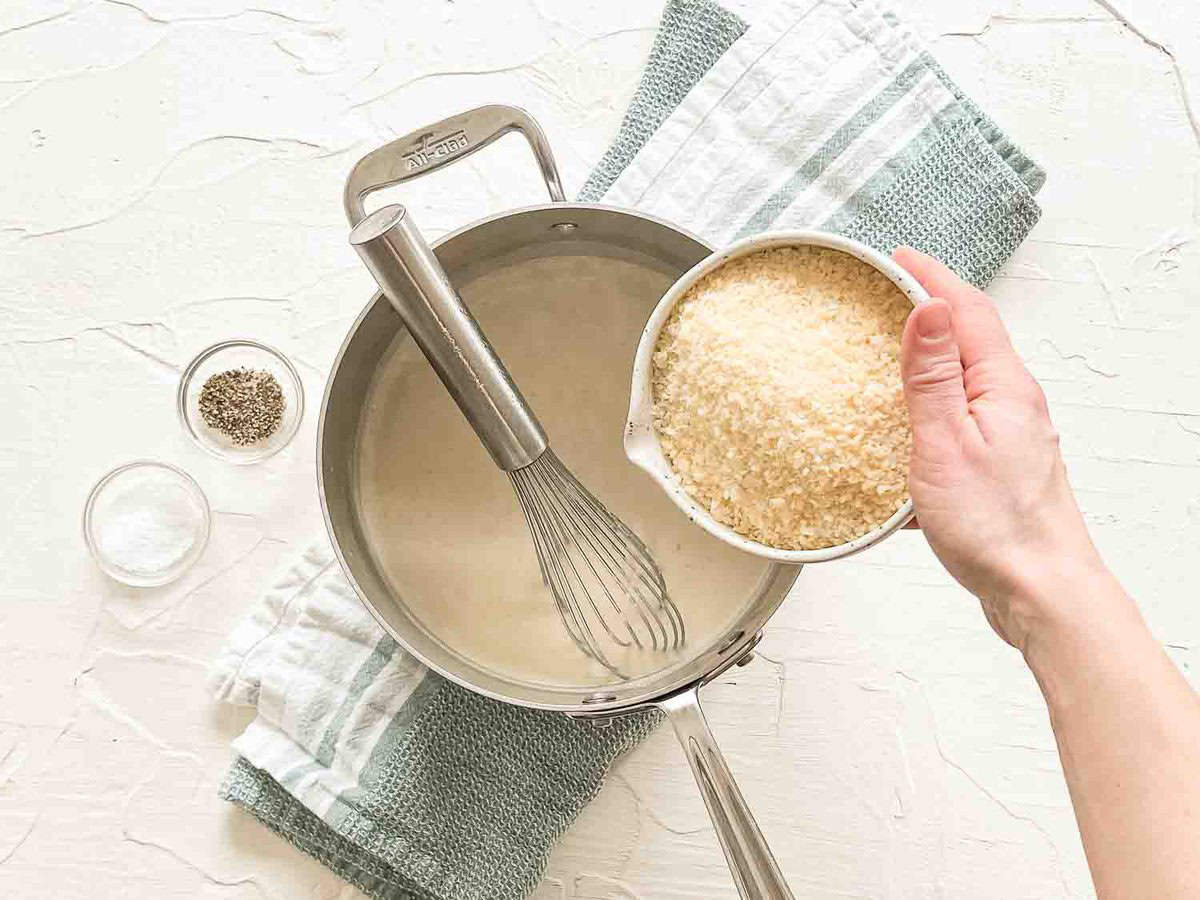 A bowl of grated parmesan cheese being dumped into a saucepan with a roux in it.