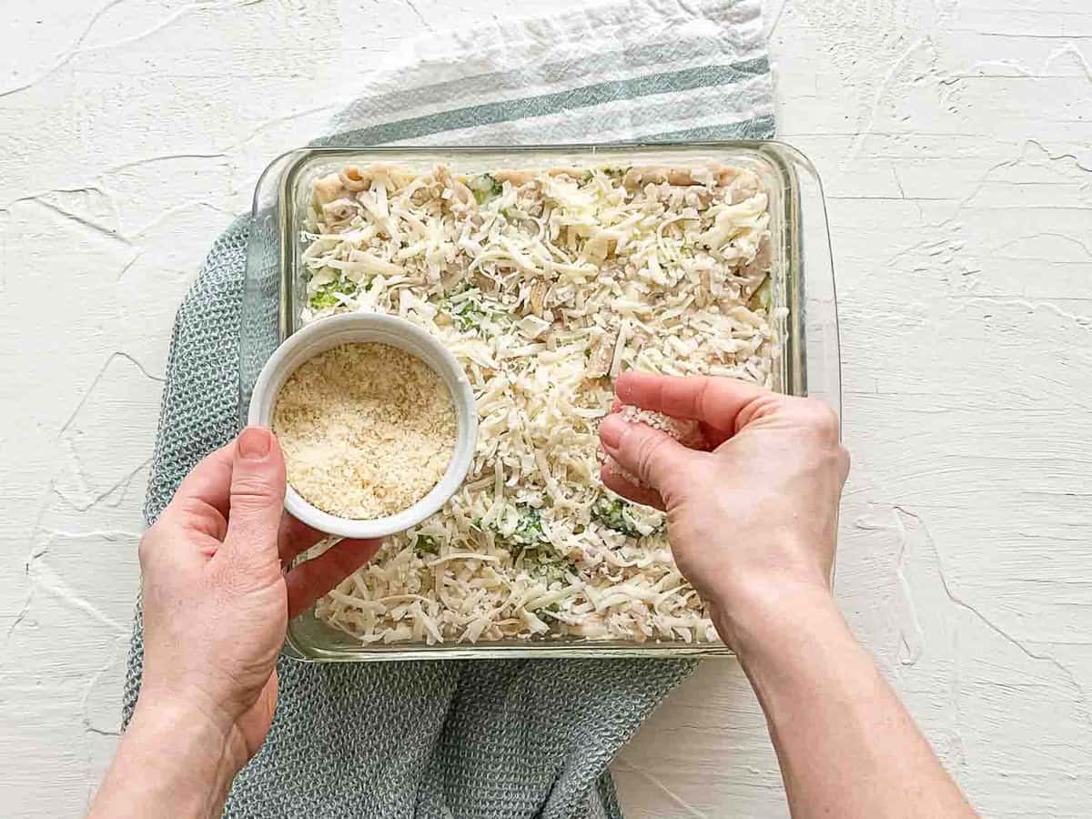 A hand sprinkling grated parmesan cheese on a baking dish with ingredients for chicken broccoli alfredo bake in it.