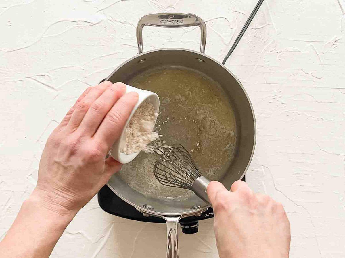 One hand dumping flour in a saucepan while the other hand is whisking the melted butter to make a roux.