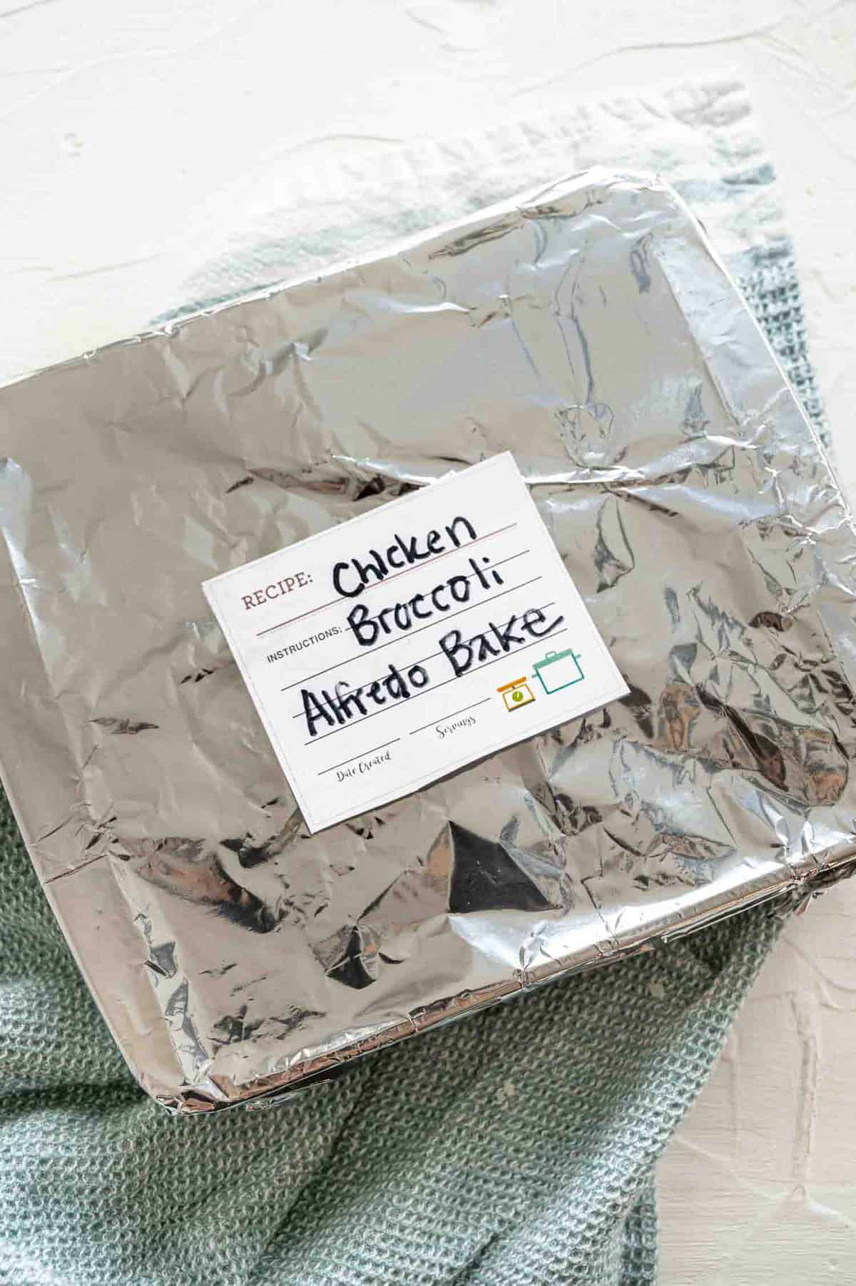 An 8x8 baking dish covered with foil and labeled chicken broccoli alfredo bake.