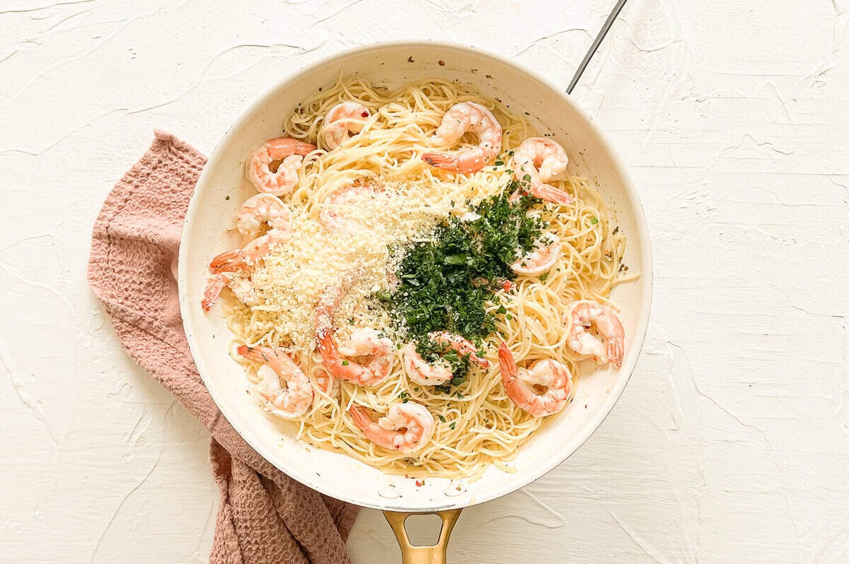 Shrimp scampi pasta ingredients in a skillet with grated Parmesan cheese and chopped fresh parsley being stirred in for the final step.