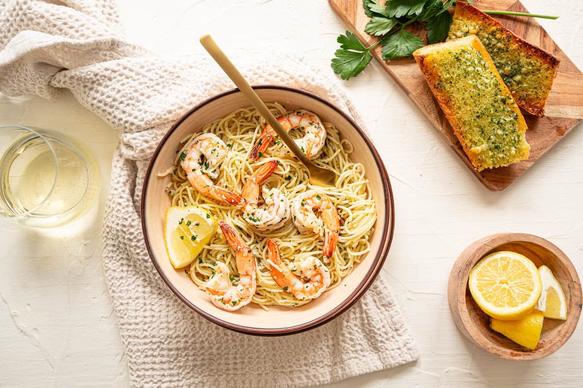 Shrimp scampi pasta in a bowl with Texas toast and lemon wedges on a cutting board next to it.