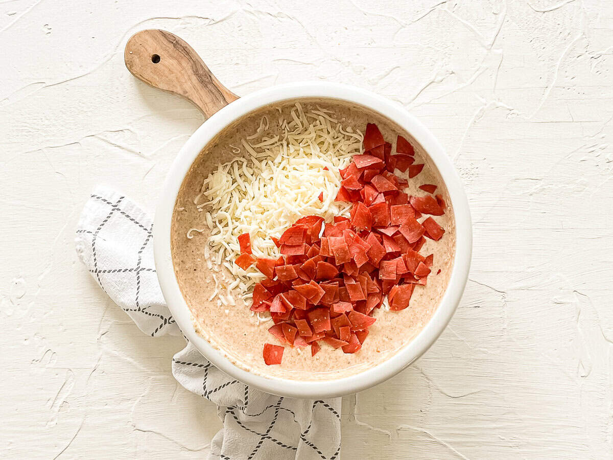Pizza muffin batter in bowl with chopped pepperoni and shredded mozzarella dumped on top to be mixed in.