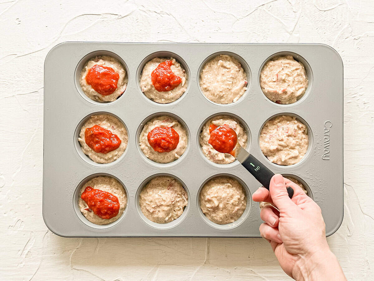 A hand using a teaspoon to put marinara sauce on top of pizza muffin batter in a muffin tin.