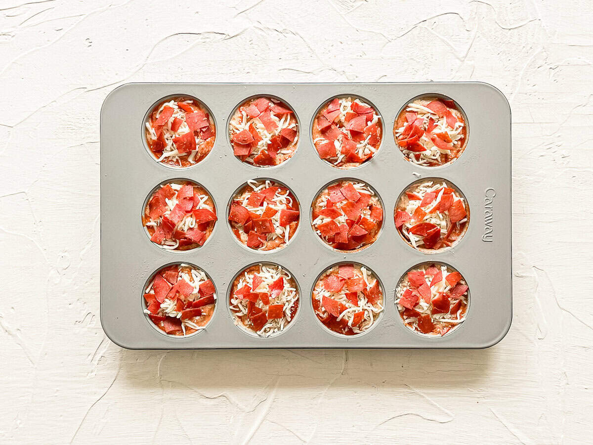 Chopped pepperoni and shredded mozzarella on top of batter in a muffin tin ready for the oven.