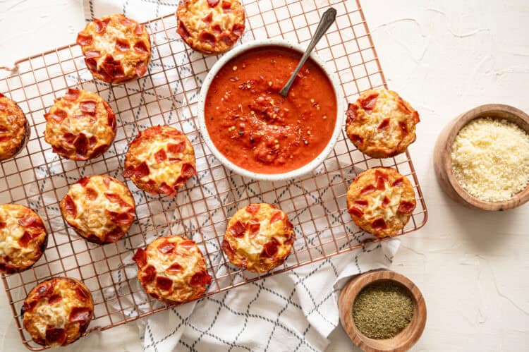 A baking rack with pepperoni pizza muffins and bowls of marinara sauce and grated Parmesan cheese.