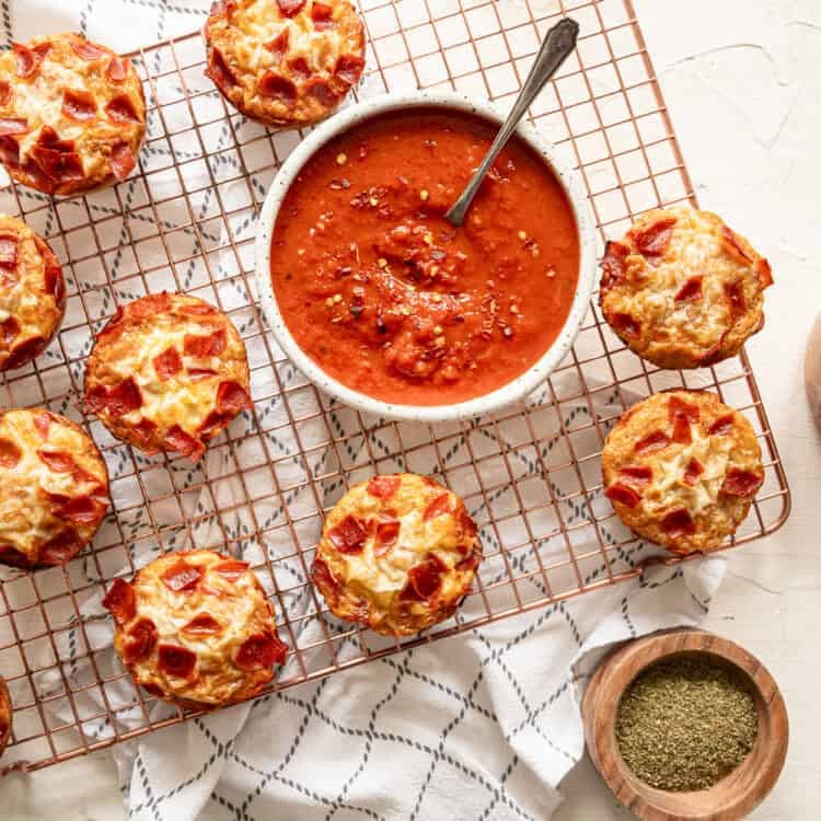 A baking rack with pepperoni pizza muffins and bowls of marinara sauce and grated Parmesan cheese.