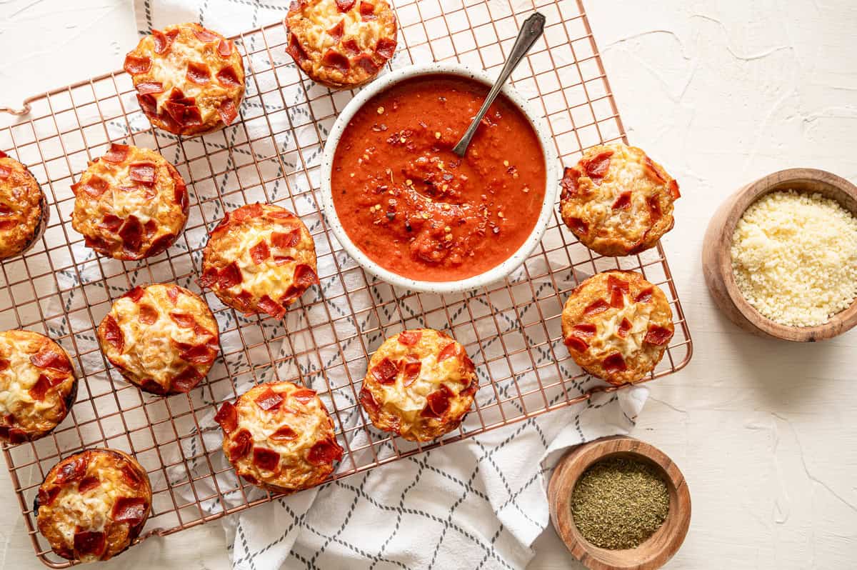 A baking rack with pepperoni pizza muffins and bowls of marinara sauce and grated Parmesan cheese.