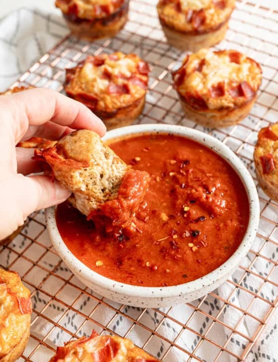 A hand dipping part of a pepperoni pizza muffin into a bowl of marinara sauce.