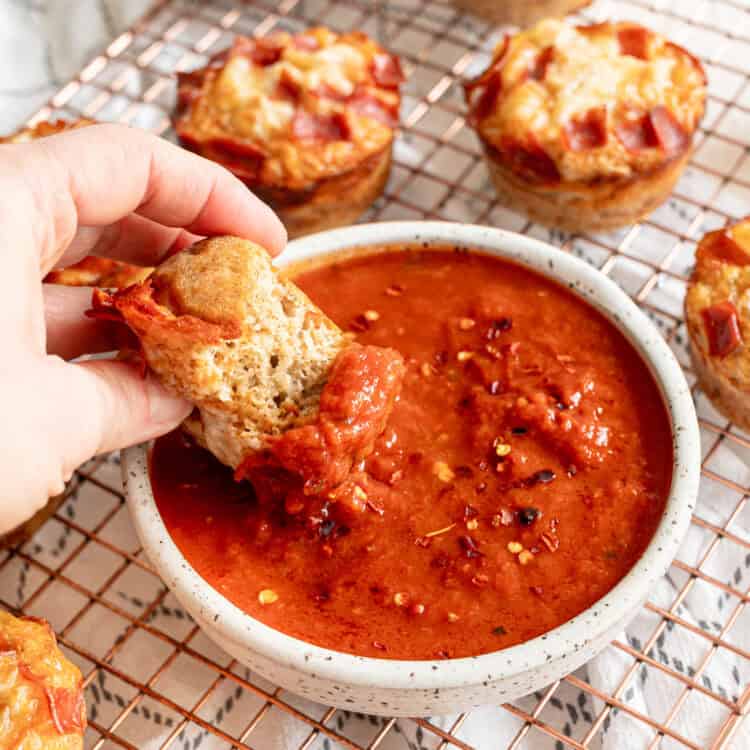 A hand dipping part of a pepperoni pizza muffin into a bowl of marinara sauce.