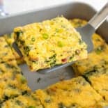 The middle slice of crustless bacon spinach quiche being lifted with a spatula out of a baking dish.