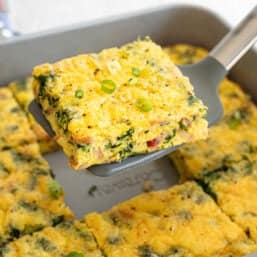 The middle slice of crustless bacon spinach quiche being lifted with a spatula out of a baking dish.