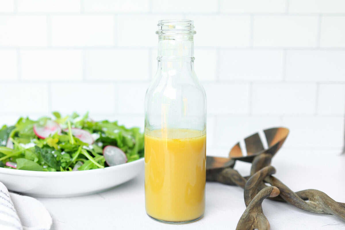 A tall thin glass bottle with golden colored salad dressing next to a salad and tongs.