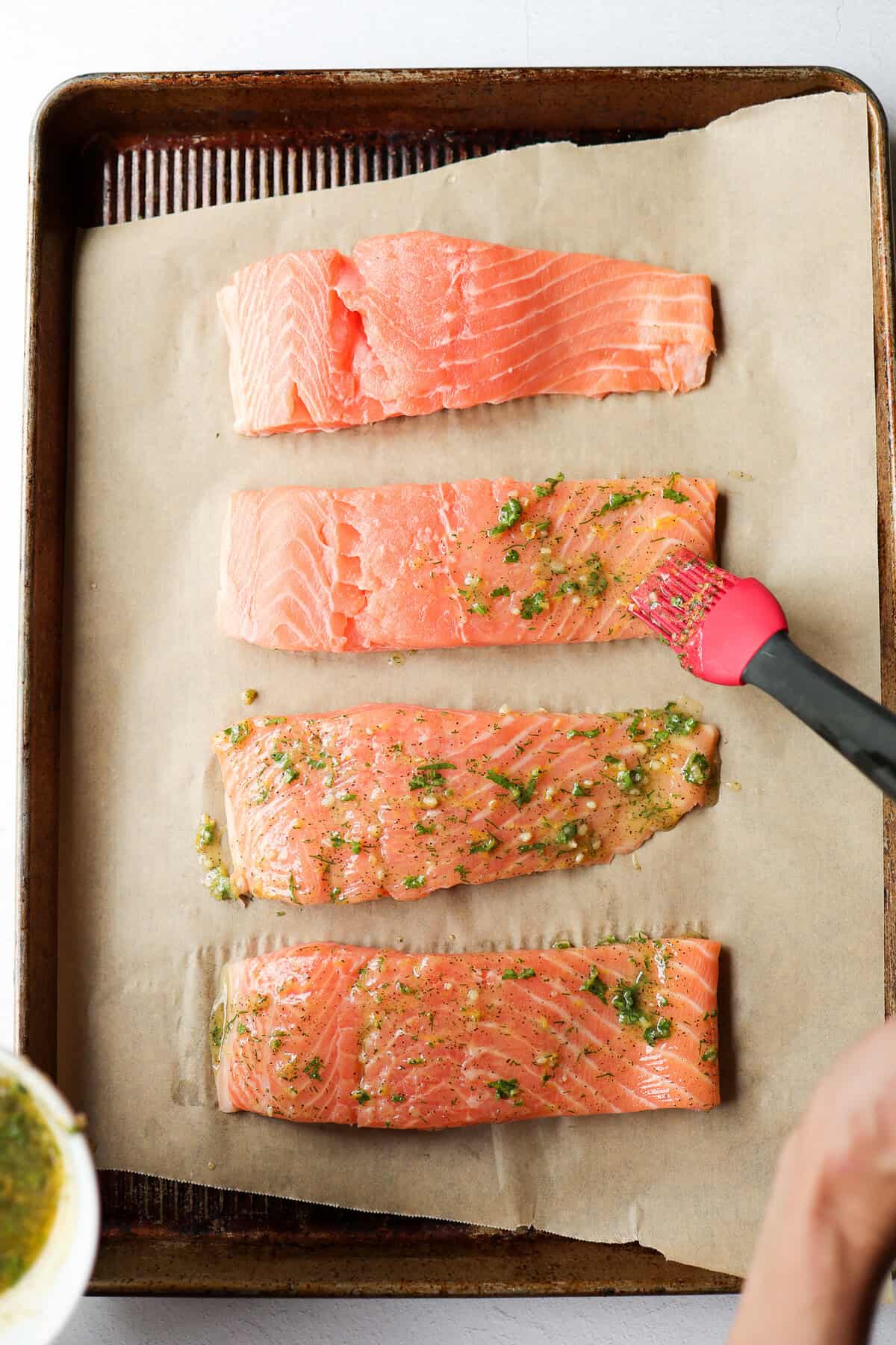 Someone brushing lemon garlic marinade on salmon fillets on a parchment-lined baking sheet.