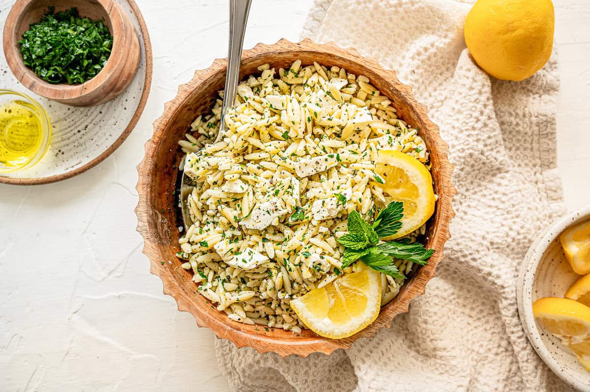 Orzo pasta in a bowl with chopped herbs, feta, and some lemon slices with a small bowl of lemons nearby.