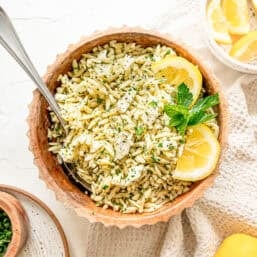 Orzo pasta in a bowl with chopped herbs, feta, and some lemon slices with a small bowl of lemons nearby.