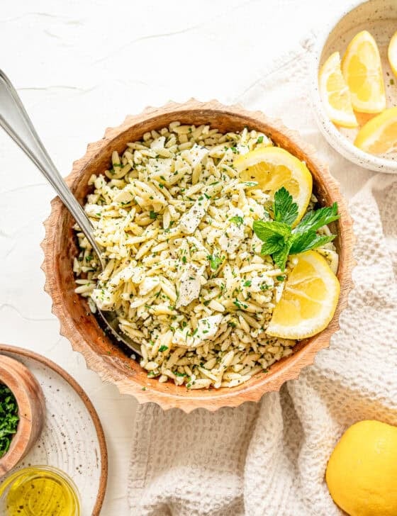 Orzo pasta in a bowl with chopped herbs, feta, and some lemon slices with a small bowl of lemons nearby.