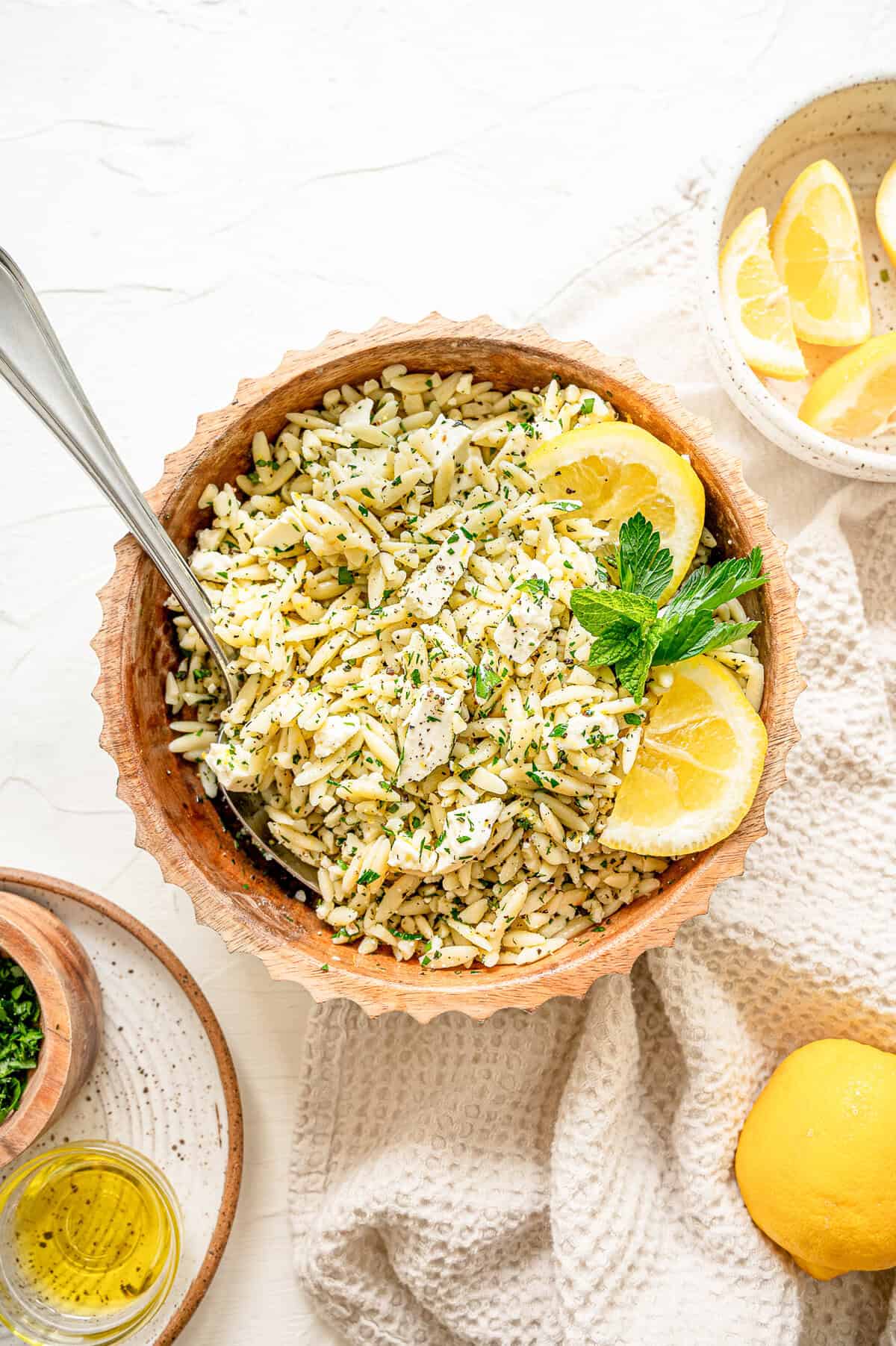 Orzo pasta in a bowl with chopped herbs, feta, and some lemon slices with a small bowl of lemons nearby.