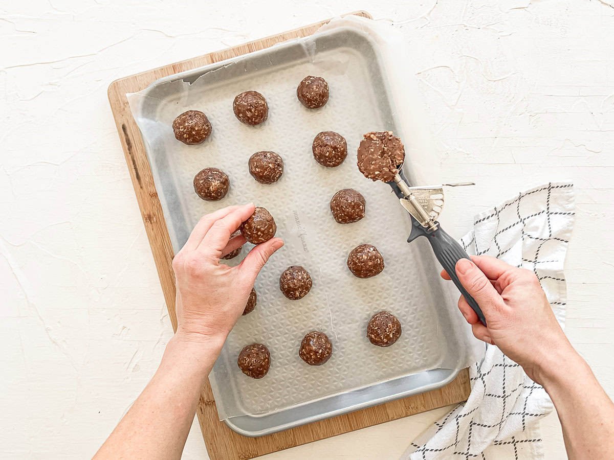 A person placing one protein ball on a baking sheet full of them with a scoop of the chocolate peanut butter mixture in the other hand ready to roll into a ball.