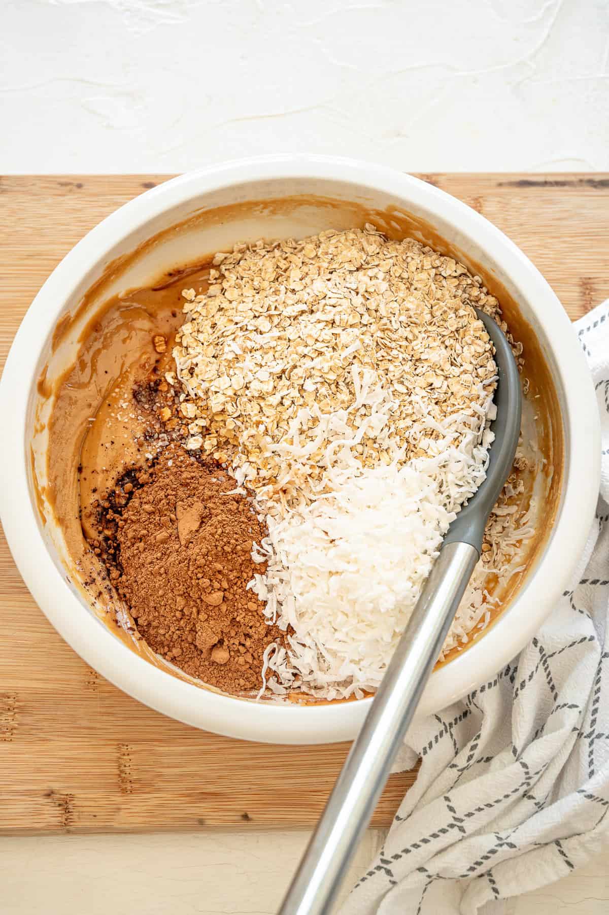 Coconut flakes, cocoa powder, and quick oats dumped on top of honey and peanut butter in a bowl.
