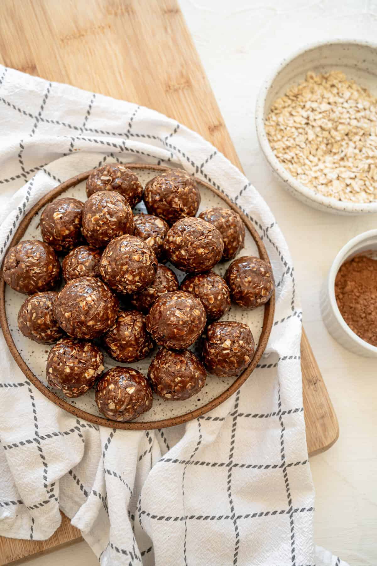 Chocolate peanut butter oatmeal balls stacked on a plate with bowls of quick oats and cocoa powder next to them.