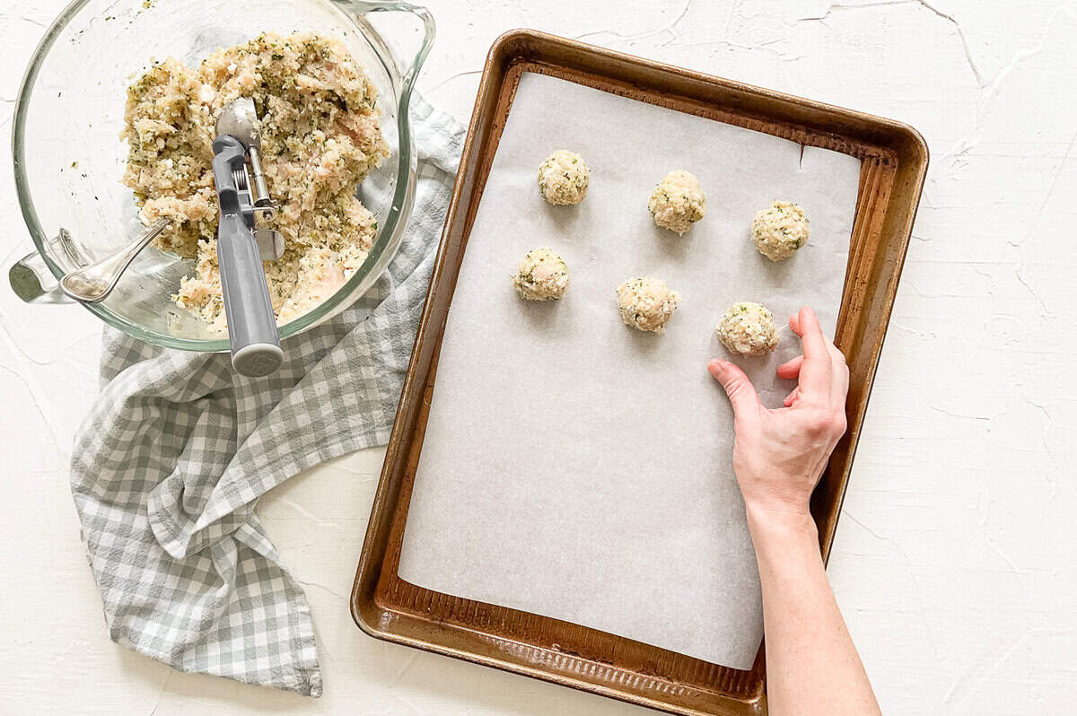 A bowl of ground meatball mixture next to a sheet pan with a few meatballs that have been rolled.