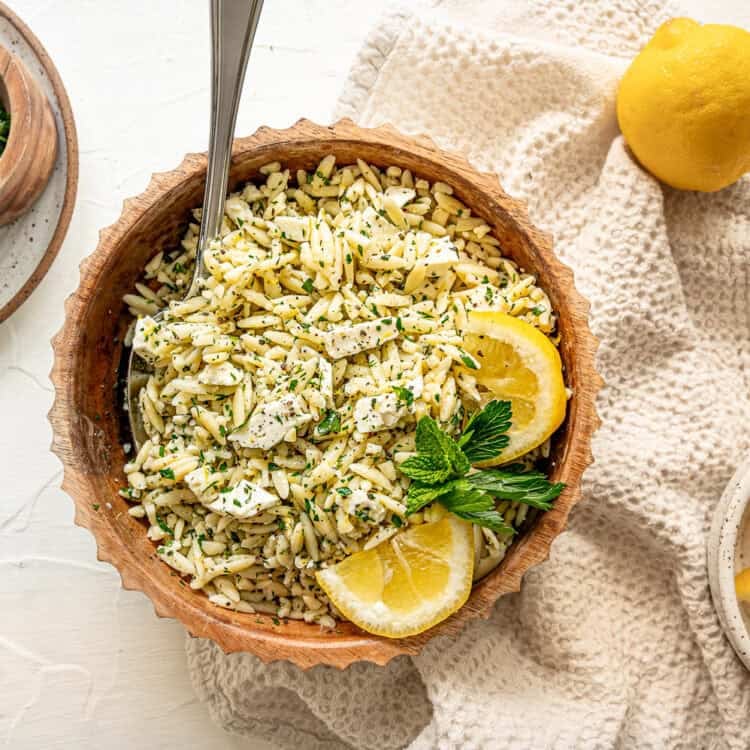 Orzo pasta in a bowl with chopped herbs, feta, and some lemon slices with a small bowl of lemons nearby.