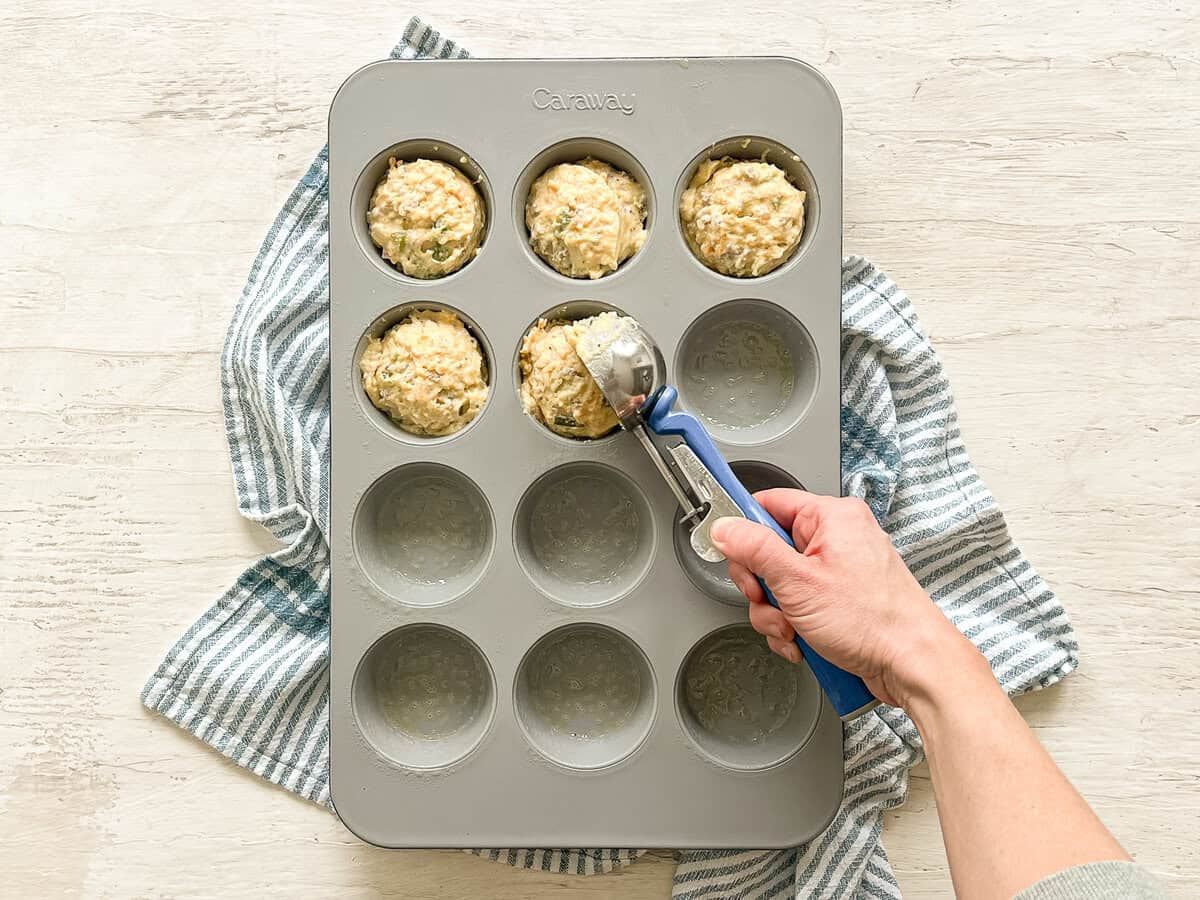 A hand using an ice cream scoop to place muffin batter in a greased muffin tin.
