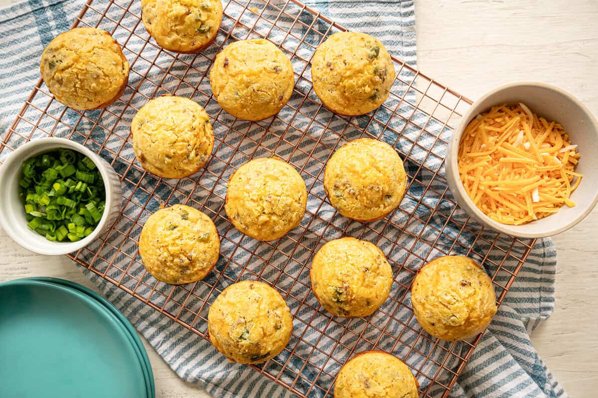 Cornmeal muffins on a cooling rack with bowls of cheddar cheese and chopped green pepper next to them.