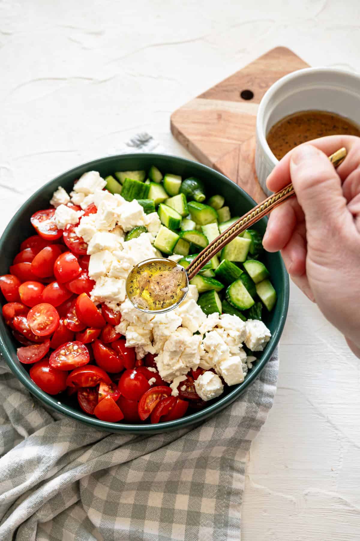 A bowl with cherry tomatoes, feta chunks, and diced cucumbers with a hand pouring a ladle of vinaigrette on it.