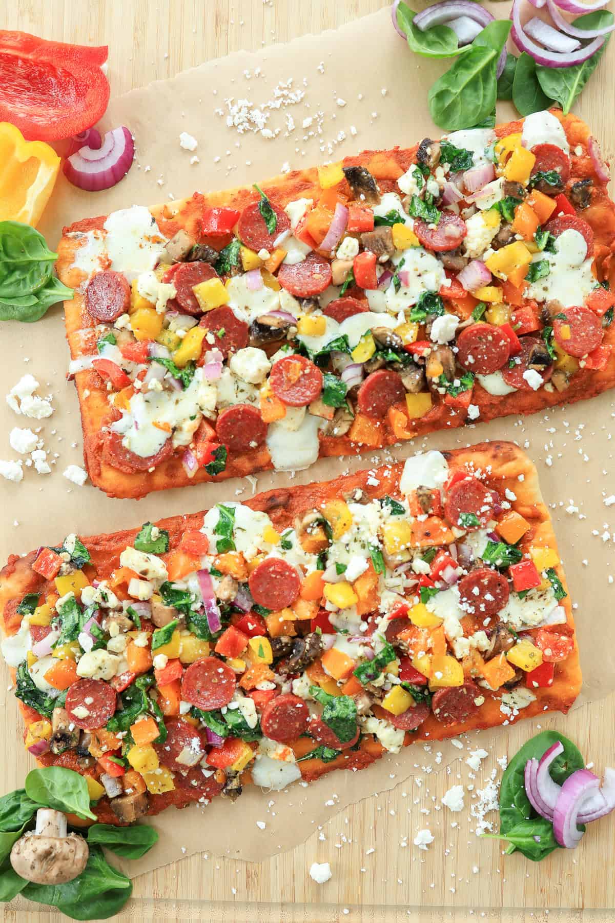 Two veggie and pepperoni flatbread pizzas not yet sliced on a cutting board.