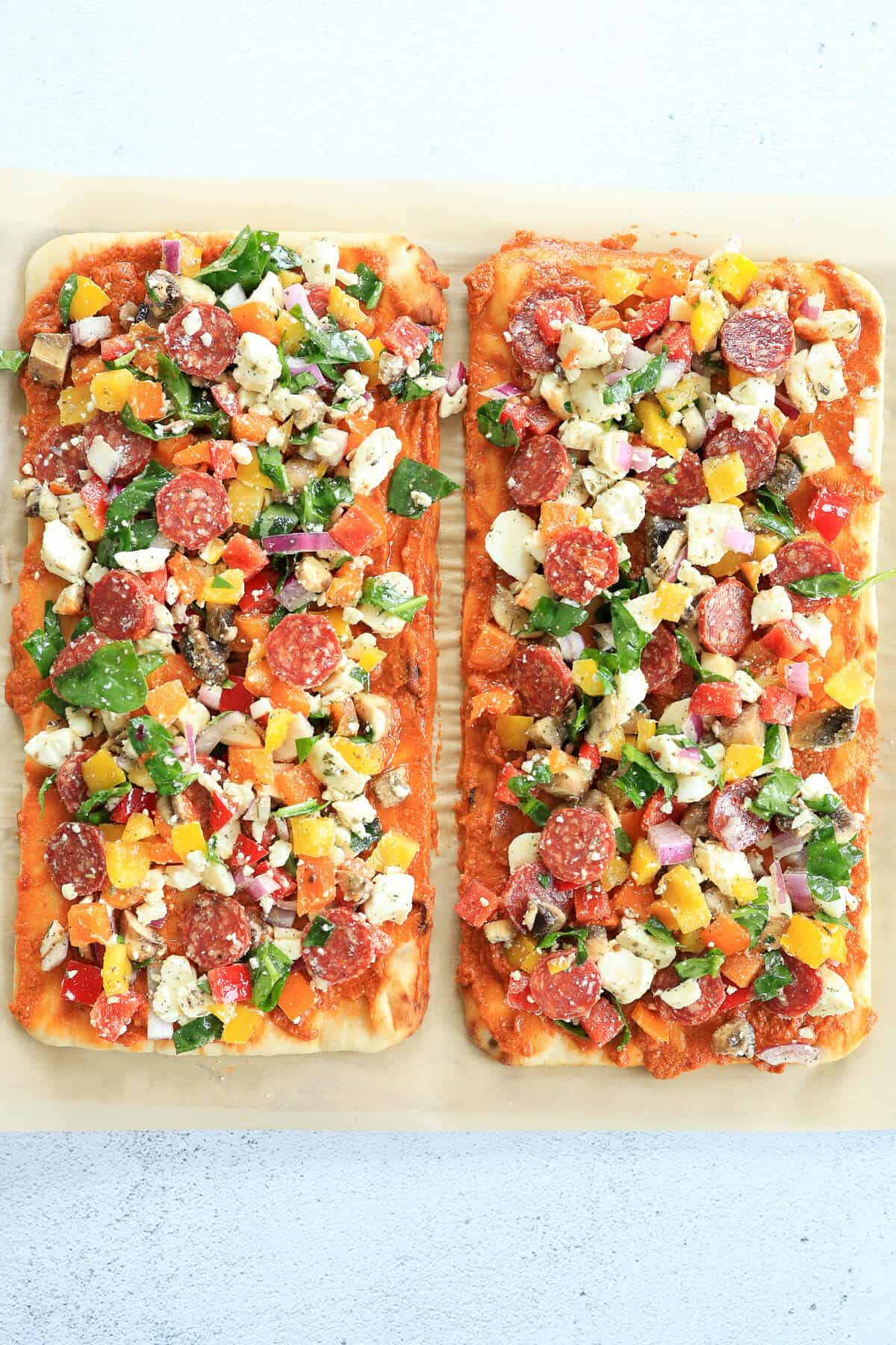 Two flatbreads covered in pizza sauce, veggies, pepperoni, mozzarella pearls, and feta cheese before being baked.