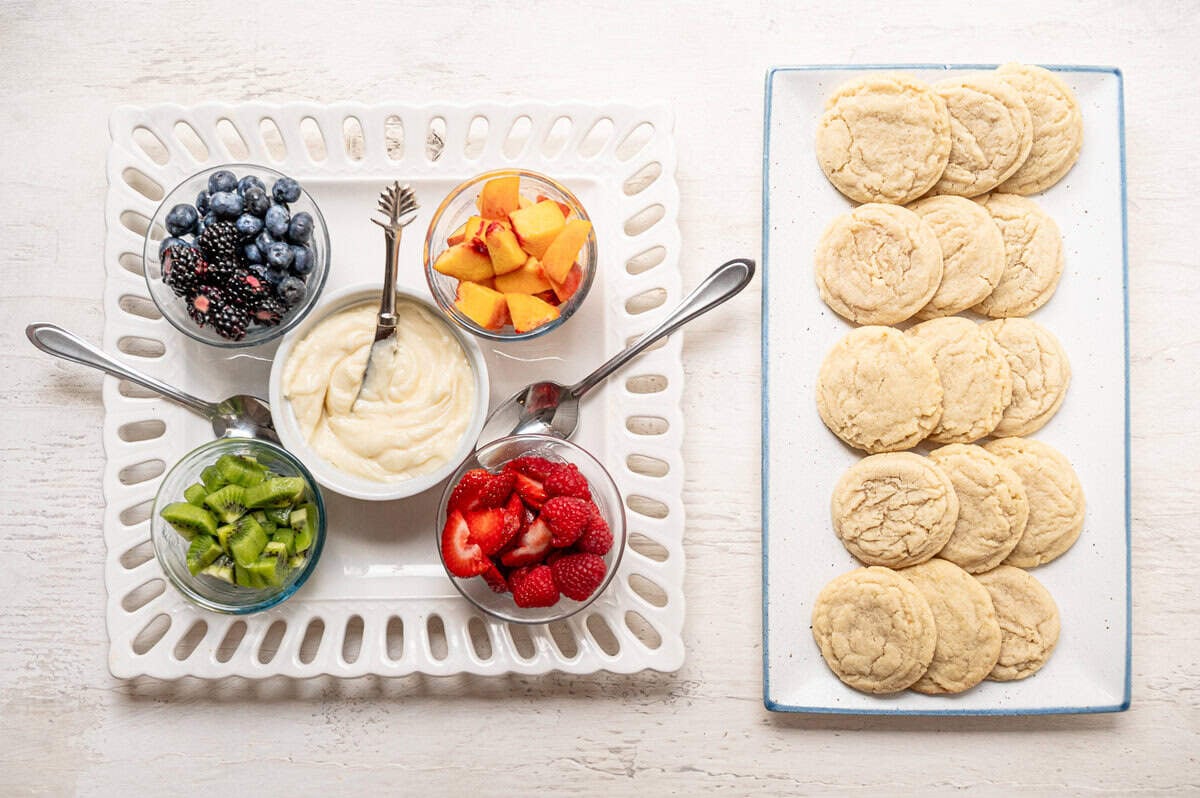 A platter of sugar cookies next to a platter with bowls of fruit and cream cheese icing.