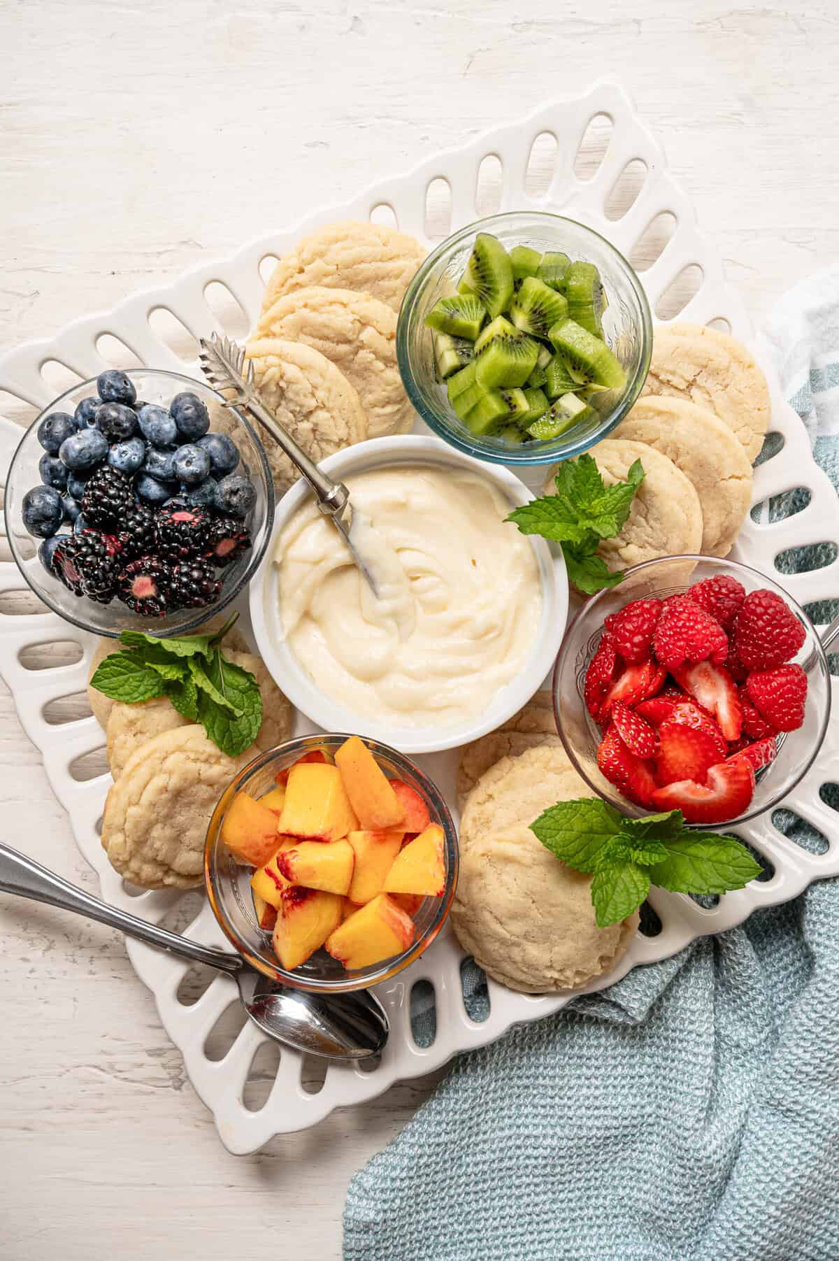 A tray for people to make fruit pizza cookies with bowls of different fruits, a bowl of cream cheese frosting, and plain sugar cookies.