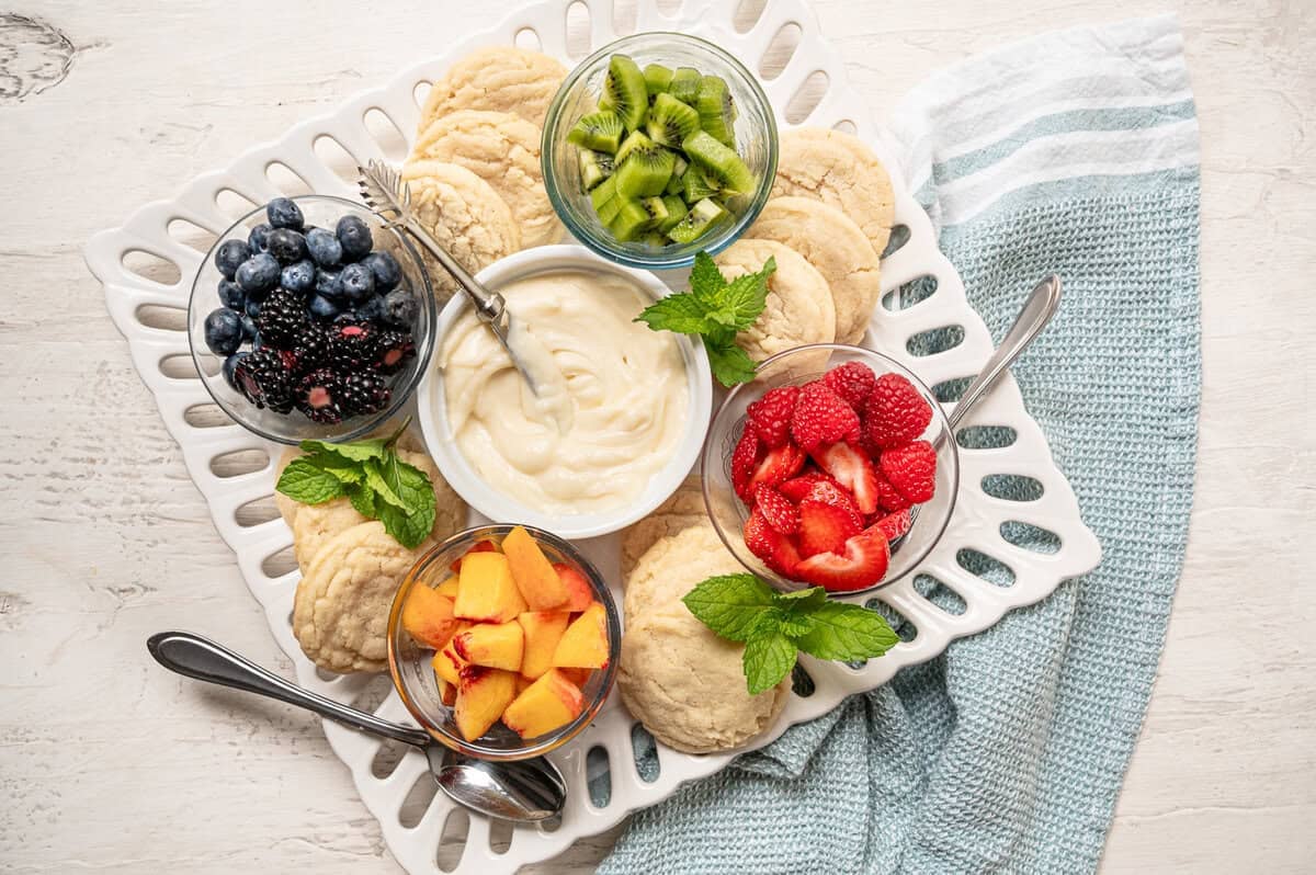 A platter for people to make their own fruit pizza cookies with bowls of fruit, and cream cheese frosting as well as plain sugar cookies.