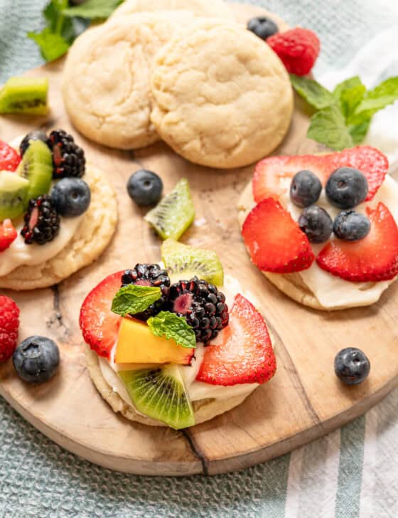 A wooden cutting board with plain sugar cookies as well as some with cream cheese frosting and fresh fruit on top.