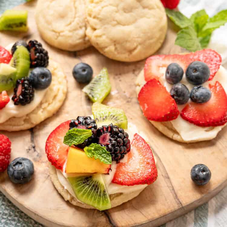 A wooden cutting board with plain sugar cookies as well as some with cream cheese frosting and fresh fruit on top.