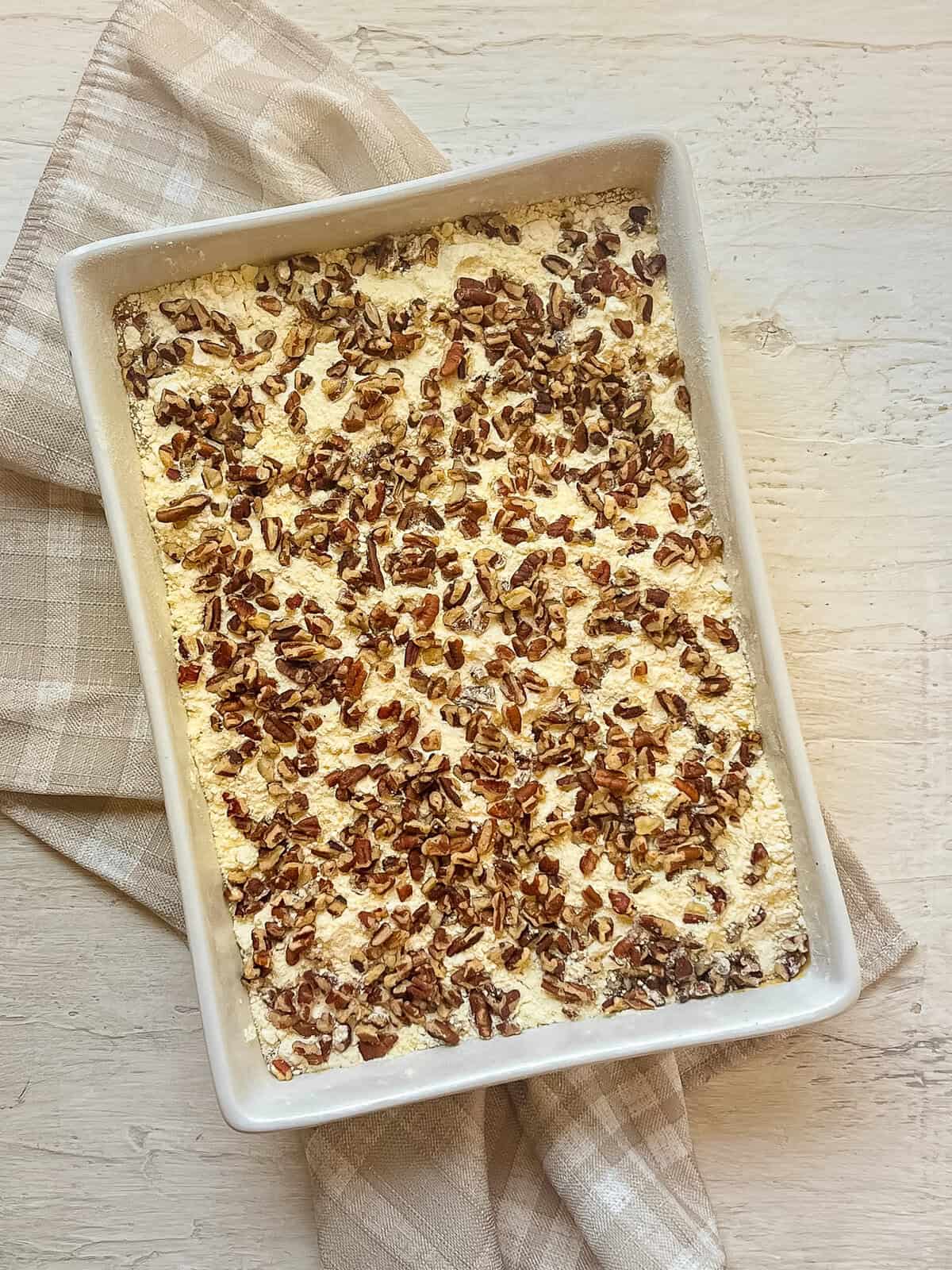 Chopped pecans sprinkled over yellow cake mix on top of wet ingredients for dump cake in a casserole dish.